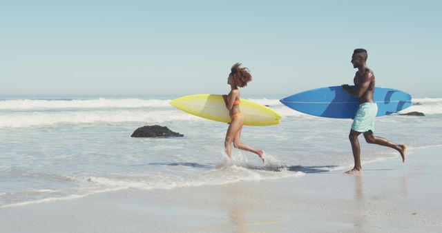 Happy diverse couple carrying surfboards on sunny beach running into the sea. Summer, hobbies, romance and vacations.