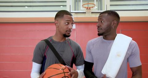 Two male teammates holding basketball and talking on indoor court under hoop