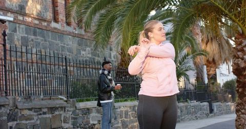 Morning urban workout stretching in pink jacket on palm-lined sidewalk holding coffee cup