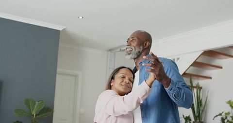 Mature couple dancing slowly in modern living room holding hands during romantic moment