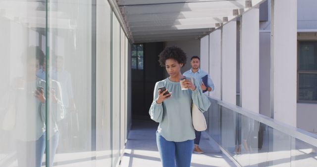 This image shows an African American businesswoman intently looking at her phone while holding a coffee cup, walking through a modern office walkway. It can be used to depict themes of mobile communication, work-life balance, professional environments, and business activities. Suitable for corporate websites, business articles, and promotional materials focusing on modern office culture.