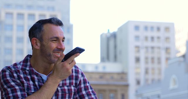 Man using a smartphone voice assistant while in an urban environment with city buildings in the background. Ideal for settings relevant to technology, communication, modern living, and urban life.
