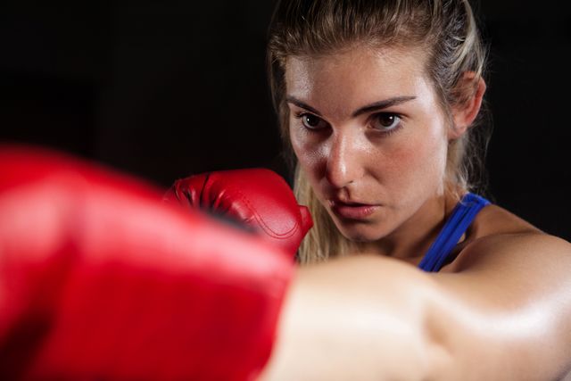 Close-up of a determined woman practicing boxing, wearing red gloves and blue top. Captures intense focus and strength. Useful for promoting fitness programs, sports gear, motivational content, and women's empowerment themes.