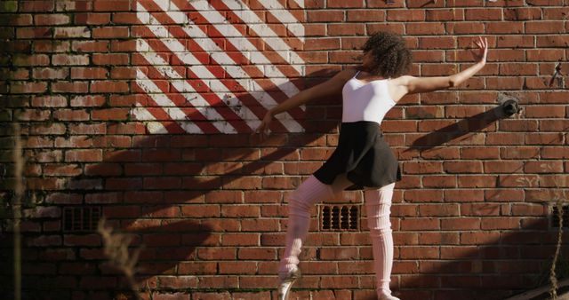 Ballet dancer gracefully posing against a brick wall with decorative painted stripes. Dress and physicality highlight artistic expression in urban settings. Ideal for uses concerning dance, art, urban culture, performances, fitness, lifestyle, and creative inspiration.