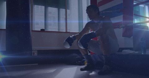 Moody boxer resting on ring apron with gloves and sunlight through gym windows