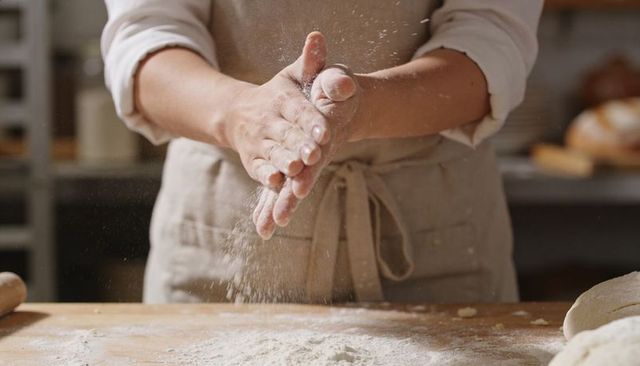 Clapping flour-covered hands scattering flour over dough on rustic wooden countertop closeup