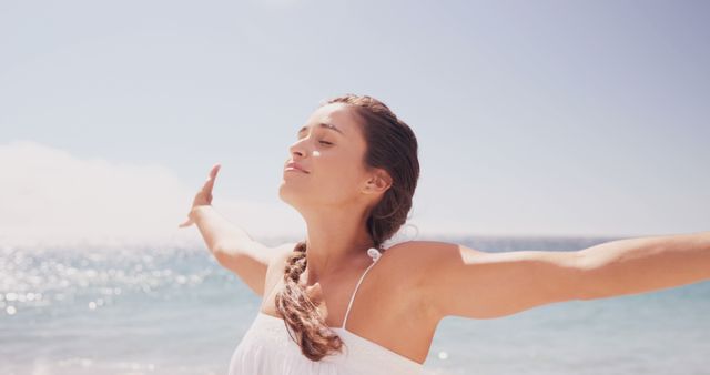 This image shows a woman with braided hair standing on a sunny beach, with her eyes closed, basking in the sunlight and fresh sea breeze. Perfect for use in travel brochures, wellness blogs, advertisements promoting relaxation and vacation destinations, or social media campaigns centered around mindfulness and living in the moment.