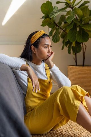Pensive asian woman on sofa wearing mustard overalls and yellow headband in sunlit minimalist room