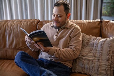 Mature asian man reading endless sunsets on leather couch, cozy home relaxing moment