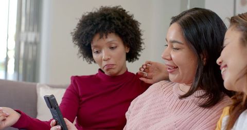 Multicultural women laughing on cozy sofa while sharing remote and enjoying tv night