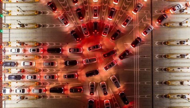 Aerial toll plaza at night showing converging cars, red taillights, symmetric lane patterns