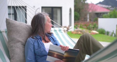 Mature woman relaxing in hammock reading sunset brochure on modern patio backyard retreat