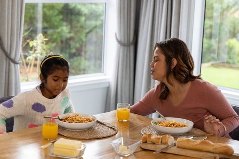 Mother leaning toward daughter sharing pasta and orange juice at sunlit home dining table