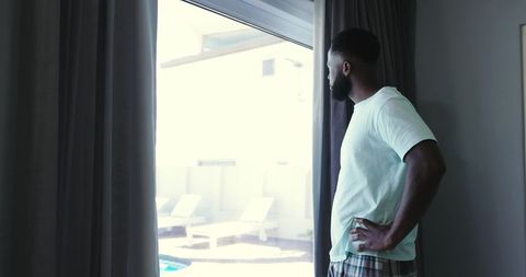 African American man standing at sliding glass door looking at sunlit pool and patio