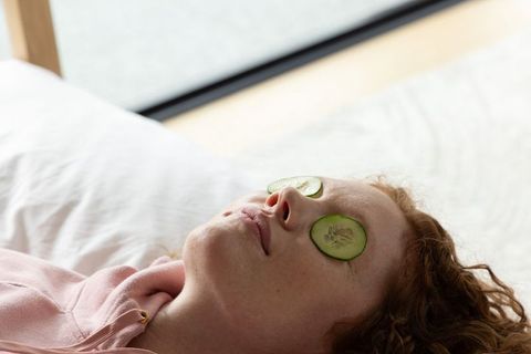 Relaxing woman with cucumber eye slices on white bed promoting self care and wellness