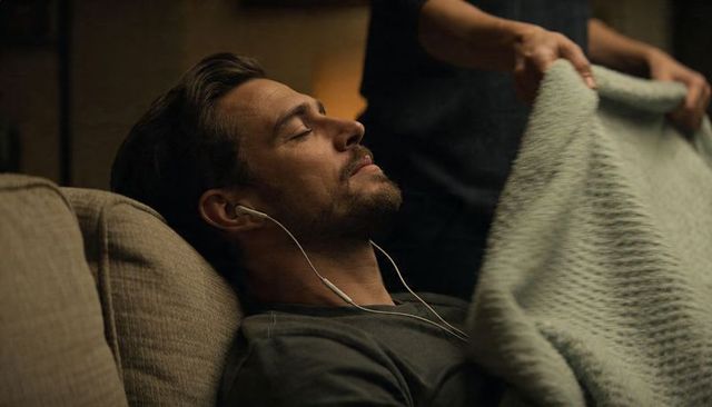 Reclining man with earbuds receiving knit blanket on sofa, cozy warm evening relaxation