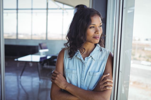 Young businesswoman standing by office window, gazing outside with a thoughtful expression. Ideal for use in professional and corporate settings, career-related articles, and business advertisements.
