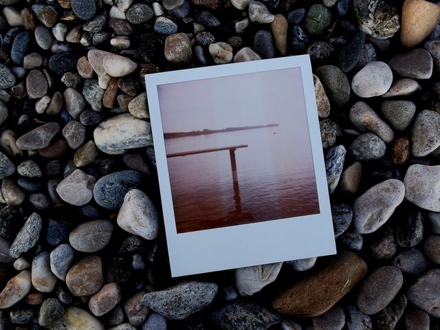 Vintage Polaroid photo featuring a wooden dock on a rocky beach foregrounded by smooth stones and pebbles. Calm water of lake or river shown in photo. Perfect for themes of nostalgia, travel memories, summer vibes, retro photography, and serene landscapes. Ideal for use in blog posts, travel websites, and social media content centered around peaceful retreats and reflection.