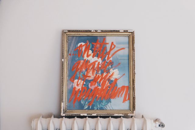 Framed painting featuring vibrant orange graffiti style text hanging on a plain white wall. Ideal for use in art gallery promotions, blog posts about contemporary art, or discussions on creative expressions and street art influences in traditional settings.
