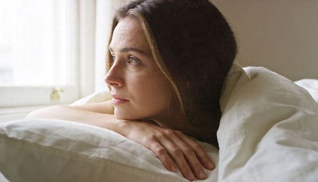 Serene woman lying in bed gazing out window, resting chin on hands in soft morning light