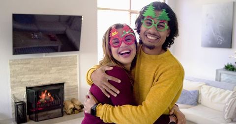 Couple Embracing and Laughing in Cozy Home Wearing Festive Novelty Glasses by Fireplace