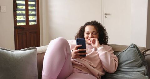 African american woman lounging on sofa scrolling smartphone in cozy pink loungewear at home