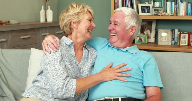 Senior couple laughing and hugging on a sofa in a cozy living room. Man wearing blue polo, woman wearing polka dot shirt. Use for retirement, senior lifestyle, love, companionship, and caregiving themes in images or advertisements.