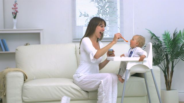 Mother is feeding her baby who is seated in a high chair. The living room setting is modern and bright, creating a cozy and familial atmosphere. Use this for visuals focused on parenting, motherhood, and home life themes. Great for blogs, brochures, and family-related advertisements.