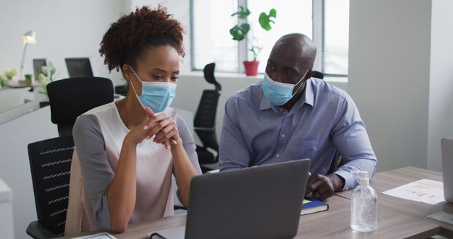 Two business professionals, wearing face masks, are collaborating over a laptop in a modern office setting. They are seated at a desk with office chairs, sanitizer placed on the desk, symbolizing a safe and sanitized work environment. This image is ideal for illustrating concepts related to workplace safety, professional collaboration, pandemic-conscious offices, and business planning.