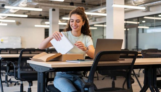 Young woman unboxing spiral notebook at modern coworking desk, smiling and working