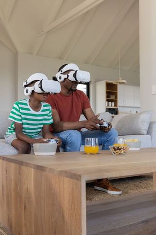 African american father and son wearing vr headsets playing games on cozy living room sofa