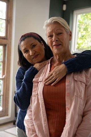Senior female partners embracing at home, showing wedding ring, sunlit cozy living room