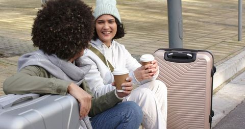 Young friends sitting on curb with luggage holding takeaway coffee, smiling and chatting