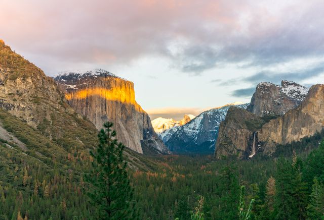 Dramatic sunset view of Yosemite National Park with sunlight illuminating rocky cliffs and snowy peaks in the distance. Dense forest covers the valley floor. Ideal for travel magazines, outdoor adventure content, and nature-inspired marketing materials.