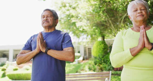Senior couple practicing yoga in an outdoor park during the day. Both individuals are in a meditative pose with hands joined in prayer. Lush greenery and bright sunlight create a tranquil atmosphere. This image can be used for promoting mindfulness, healthy lifestyles, senior wellness, fitness programs, and nature retreats.