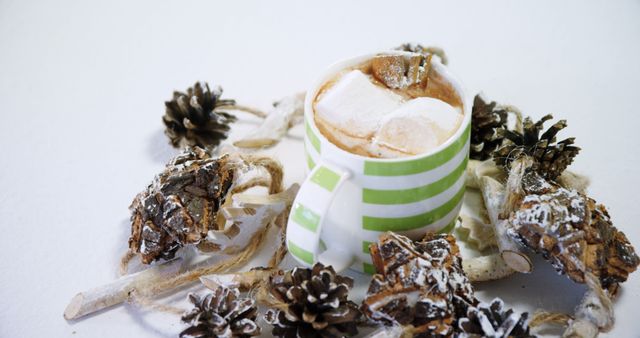 Hot chocolate with marshmallows in a green striped mug sitting among pinecones. Suitable for festive holiday advertisements, winter drink promotions, cozy lifestyle blog posts, or seasonal social media content.