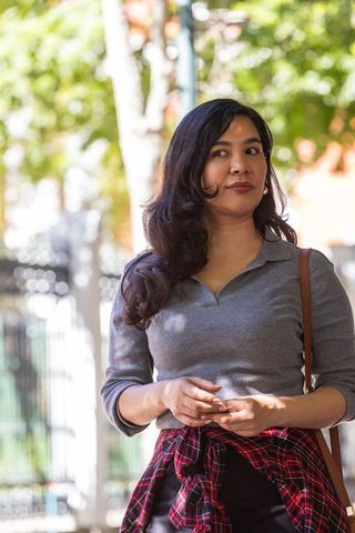 Urban woman standing on sunlit sidewalk holding smartphone looking left in plaid shirt