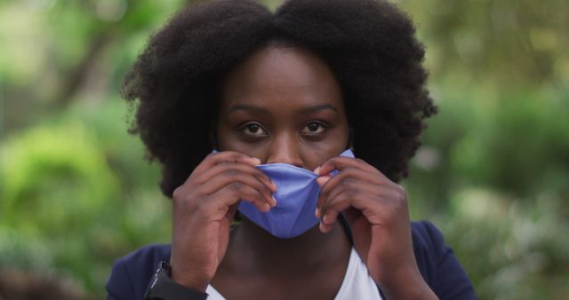 African American woman with curly hair adjusting her blue face mask while standing in an outdoor setting with greenery in the background. Ideal for use in articles or promotional materials related to health and safety, public health campaigns, pandemic prevention measures, outdoor activities during pandemic, or responsible behavior in public spaces.