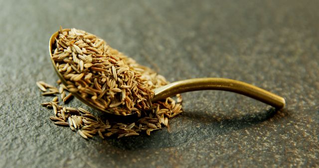 Image features aromatic cumin seeds overflowing from a gold-colored spoon placed on a textured stone surface. Perfect for use on cooking websites, food blogs, and culinary magazines to highlight ingredients, recipe articles, or spice profiles. Can also be effective for marketing gourmet food products or spice collections.