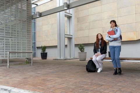 Two female students chatting and studying on campus bench with backpacks and red folder