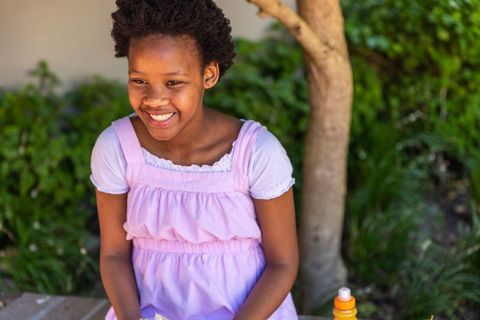 Joyful african american girl sitting on bench smiling while holding snack in purple dress