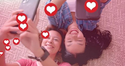 Two young women lying on rug taking selfies with smartphones and floating red like hearts