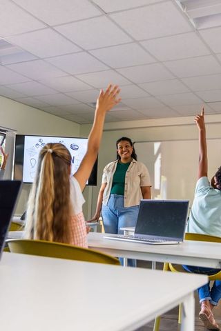 Female teacher engaging teenage students raising hands in modern tech classroom with laptops