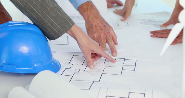 Architects and engineers are collaborating over detailed blueprint plans of a building project at an office desk. Workers' hands are pointing out specific parts of the layout, indicating active discussion and planning. A blue hard hat is present, emphasizing construction safety and professional planning. This image is ideal for illustrating teamwork, engineering projects, and architectural discussions.
