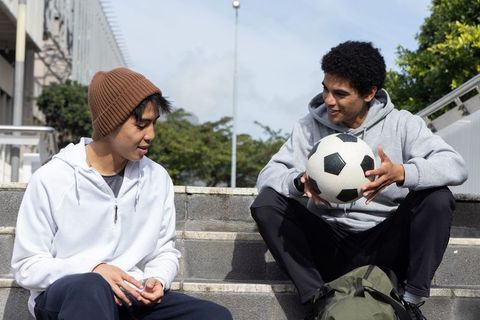 Diverse teenage friends sitting on steps sharing soccer ball and casual campus hangout