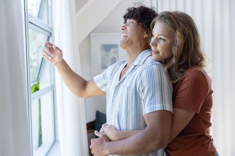 Young couple embracing by bright casement window, pointing outside, cozy home moment