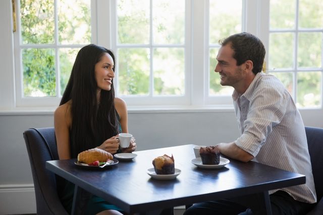 Young couple sitting at a table in a cafe, enjoying coffee and pastries while smiling and talking. Ideal for use in content related to dating, relationships, cafes, morning routines, and social interactions.