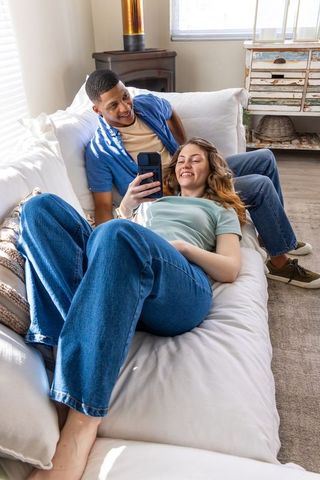 Diverse couple lounging on sunlit sofa sharing smartphone, cozy casual home relaxation