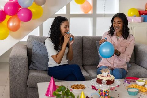 Multicultural friends inflating balloons on sofa celebrating cozy home birthday party