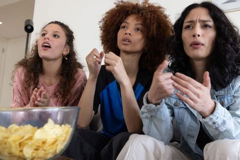 Diverse friends watching dramatic tv show on couch, reacting with suspense